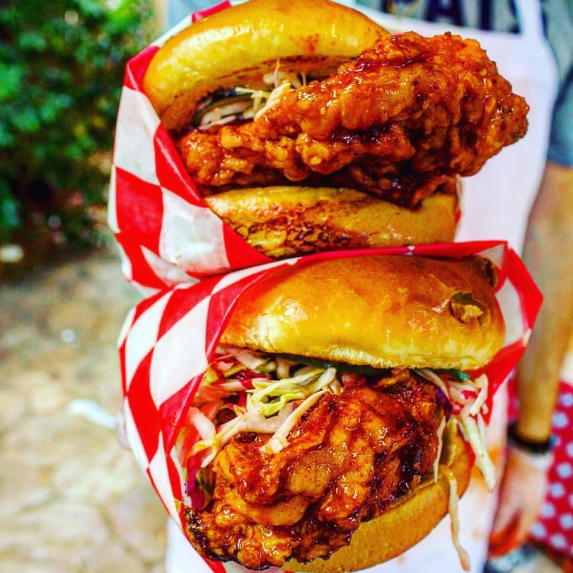Two 2 Neighbor's fried chicken sandwiches with slaw and sauce, wrapped in red and white checkered paper.