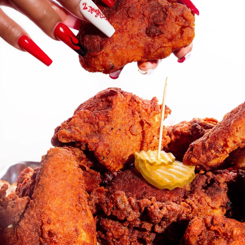 A hand with long red and white nails holds a piece of 2 Neighbor's fried chicken over a pile of chicken.